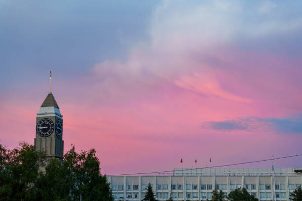 A clock tower and building are silhouetted against a vibrant pink and purple sky at sunset. The peaceful urban scene is highlighted by the dramatic colors of the evening sky, with no people in view