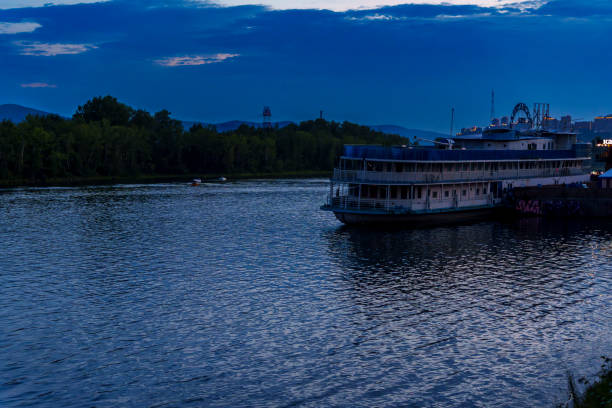 A riverboat docked on a calm river during dusk, surrounded by trees and mountains. The peaceful scene is captured during the blue hour, with reflections on the water and no people visible