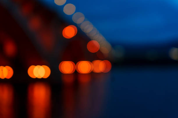 A blurred, soft-focus view of an illuminated bridge over a river at dusk, with reflections shimmering on the water. The abstract scene captures the urban structure during the blue hour