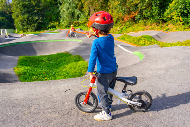 A young boy in a red helmet rides a bicycle on a paved surface. The boy is wearing a blue shirt and blue jeans