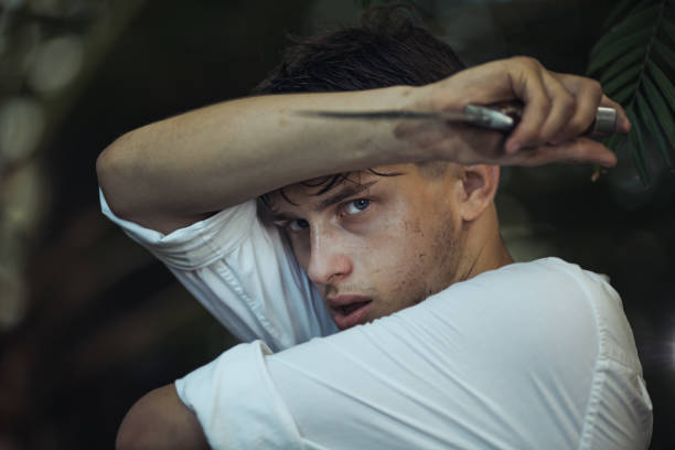 Intense young man with a knife raised defensively in a dense jungle, dirty and tired. Captures themes of danger, survival, and resilience. Ideal for high-quality promotional materials and advertisements.