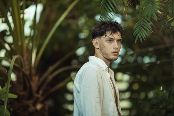 Young man with a concerned expression standing in a lush jungle, appearing lost. Wearing an open white shirt, he embodies themes of survival, exploration, and adventure. Ideal for high-quality promotional materials and advertisements.