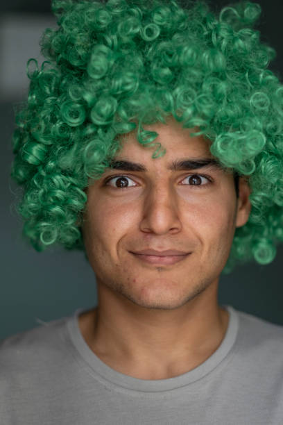 Young man wearing a bright green curly wig is making a funny face, looking directly at the camera with a slight smile and raised eyebrows
