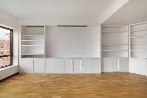 Empty built-in white cabinet on wooden laminate floor with big window in empty bright airy room in housewarming. Concept of housewarming and convenient storage.