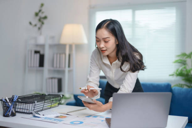 Focused young asian businesswoman using smartphone and analyzing financial charts while working with documents and laptop computer in a modern office
