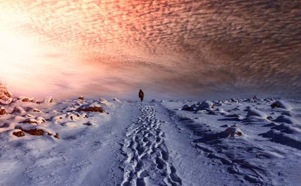 A lone traveler walks along a snow-covered path with a dramatic sunset sky, evoking feelings of solitude and adventure in a winter landscape.
