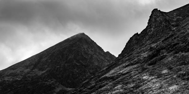 A striking mountain landscape features sharp peaks and rocky slopes under a cloudy sky. The black and white contrast highlights the rugged terrain, emphasizing the natural beauty.