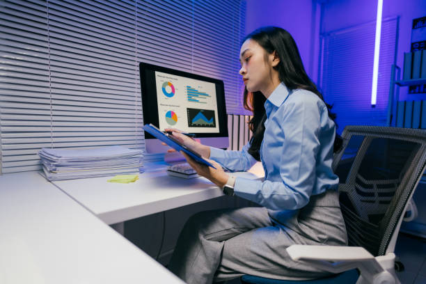 istock Young businesswoman is working late in a modern office, analyzing charts on her computer and taking notes on a clipboard, focused on her tasks in the dimly lit workspace 2185215854