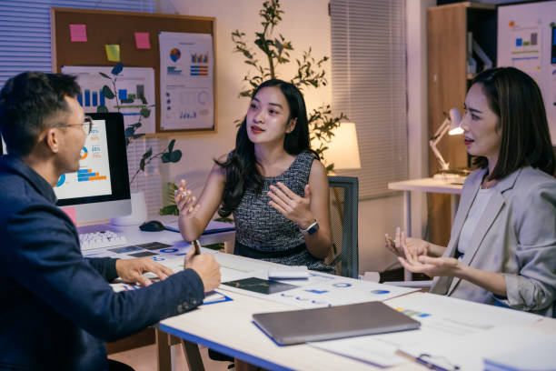 istock Three asian businesspeople are discussing strategy in a brightly lit office at night, analyzing charts and graphs on their computer and printouts 2185215995