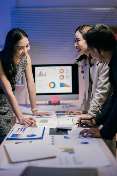 istock Four asian businesspeople are smiling and working together, analyzing charts and having a discussion in a modern office at night 2185216014