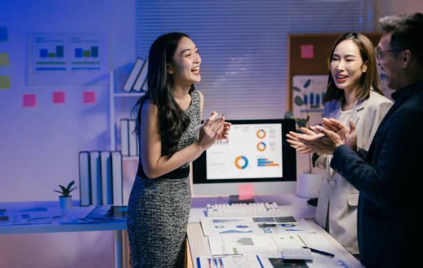 istock Three asian businesspeople clapping and laughing, satisfied with their work in a brightly lit office at night, surrounded by charts and graphs on a desk and computer screen 2185216019