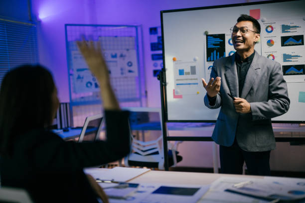 istock Asian businessman is leading a presentation in a modern office at night, explaining charts and data to his colleagues, while a businesswoman raises her hand to ask a question 2185216051