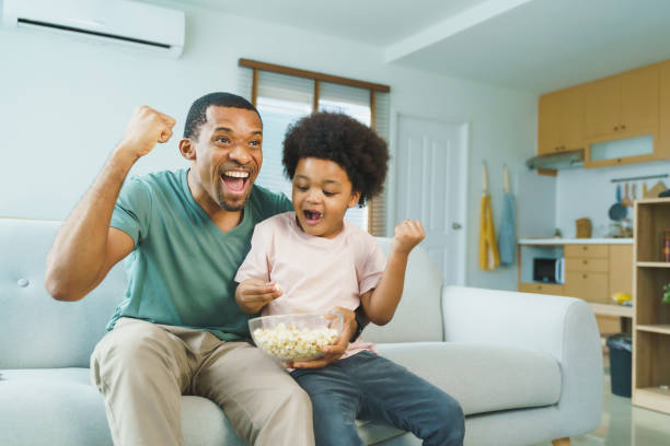 istock Black African American Father and son enthusiastically watching TV while eating popcorn on a living room sofa 2185221073