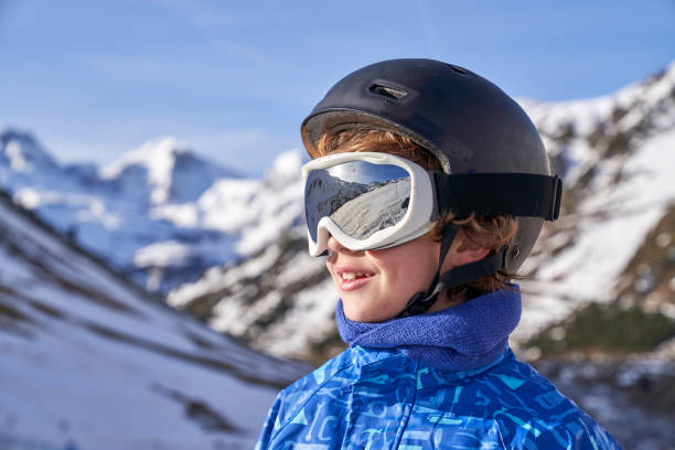 Positive kid in protective goggles and helmet smiling and looking away on blurred background of snowy mountains on sunny day on ski resort