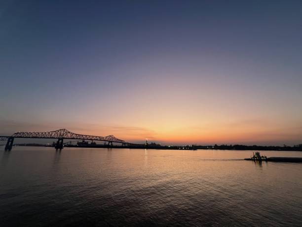 A serene sunset over a calm Mississippi River, with the silhouette of a bridge and distant city skyline against a gradient sky transitioning from orange to blue. The reflection on the water adds to the peaceful ambiance.