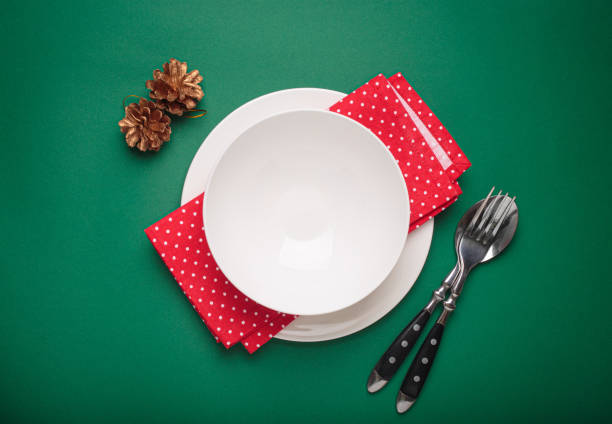Empty white plate, silverware, a red napkin with white polka dots, and golden pine cones are creating a festive christmas table setting on a vibrant green background, ready for a holiday meal