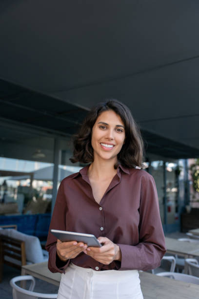 Vertical portrait of middle eastern businesswoman using tablet pc app standing at office business building outdoors. Smiling indian or latin hispanic woman holding digital computer, looking at camera
