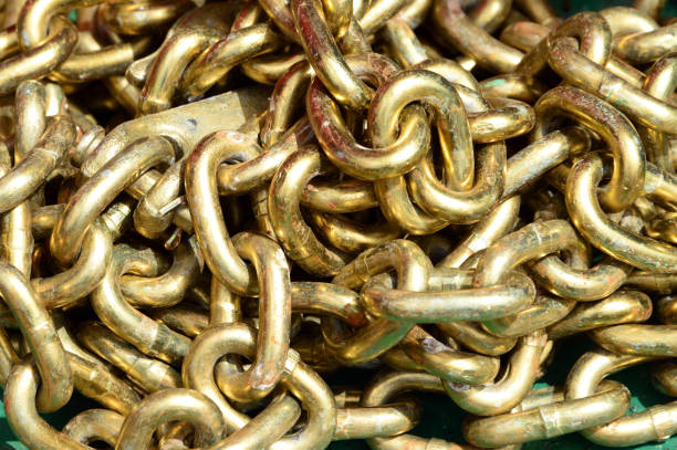 Chain background. Heavy-duty metal chain links with a gold colored finish. The links are thick and industrial in nature, arranged in an overlapping pattern that fills the entire frame. The lighting in the photograph highlights the curved surfaces of the links, creating a mix of bright reflections and shadows that emphasize the three-dimensional nature of the chain structure.