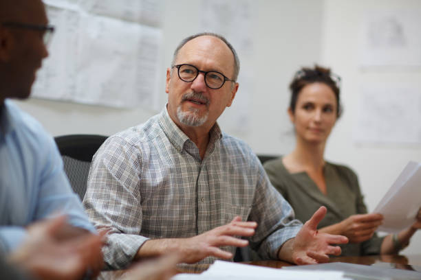 istock Senior man talks serious explaining in meeting with work colleagues while sitting at table in design office 2185295883
