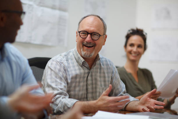 istock Senior man talks smiles laughs in meeting with work colleagues while sitting at table in design office 2185295904