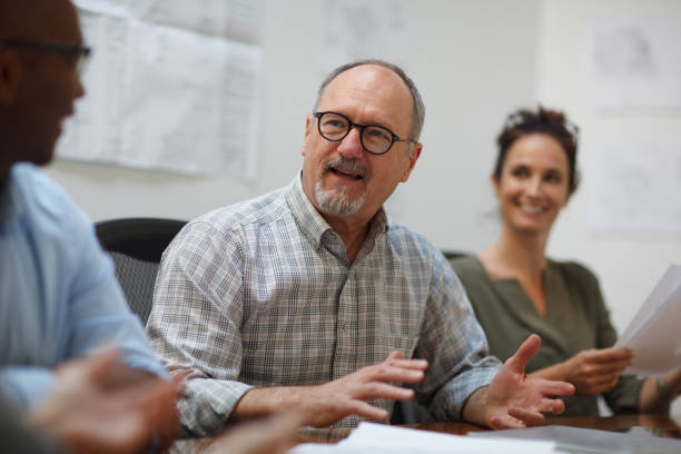 istock Senior man talks explaining in meeting with work colleagues while sitting at table in design office 2185295912