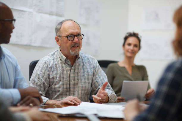 istock Senior man talks serious explaining in meeting with work colleagues while sitting at table in design office 2185295925