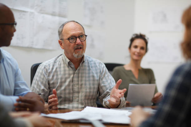 istock Senior man talks serious explaining in meeting with work colleagues while sitting at table in design office 2185295945