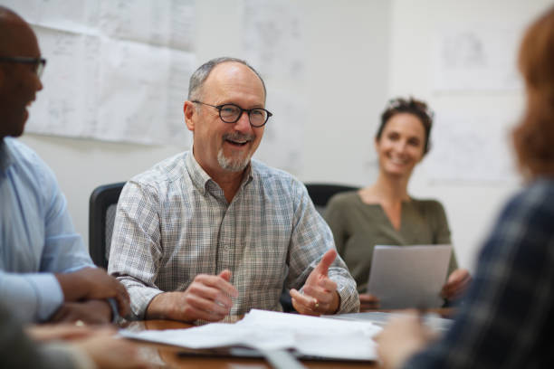 istock Senior man talks smiles laughs in meeting with work colleagues while sitting at table in design office 2185295968