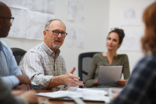 istock Senior man talks serious explaining in meeting with work colleagues while sitting at table in design office 2185296040