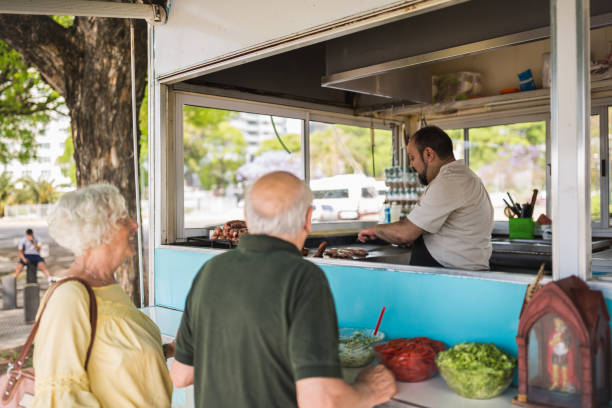 A couple is being served at a food truck during midday. The vendor is seen preparing food while the couple waits at the counter, with fresh ingredients visible.