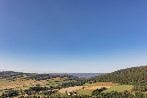 A serene rural view near Sarny, Lower Silesia, showcasing vast blue skies, rolling green hills, and patchwork fields dotted with scattered homes and a communication tower in the distance. A tranquil escape into nature.