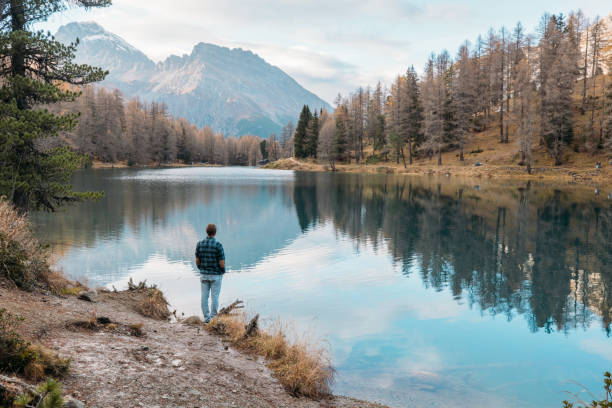A man stands alone, reflecting by a calm alpine lake framed by autumn trees and majestic mountains, embracing the peace and solitude of nature.