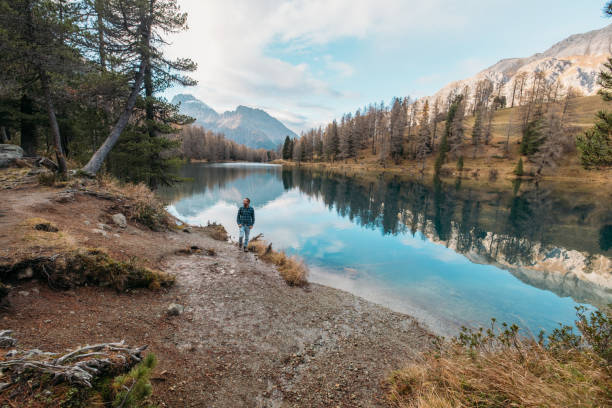 A man strolls along the shore of a serene alpine lake, surrounded by autumn trees and majestic mountain views, enjoying the tranquility of nature.