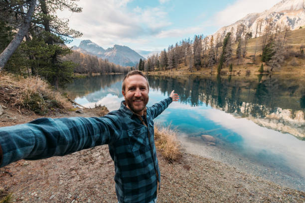 A cheerful man in a plaid shirt takes a selfie with a stunning mountain lake and autumnal scenery in the background, reflecting clear skies and rugged peaks.