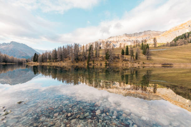 A tranquil alpine lake with crystal-clear water reflecting rugged peaks, autumn trees, and a vibrant blue sky under soft clouds, showcasing pristine natural beauty.