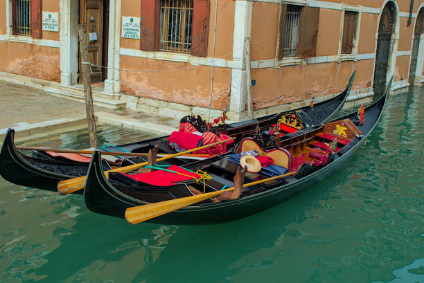 Venice, Italy - October 15, 2024: Two Venetian gondolas are moored next to a weathered building waiting for tourists. Luxurious red and black seats, intricate gold accents and a hat on one seat.