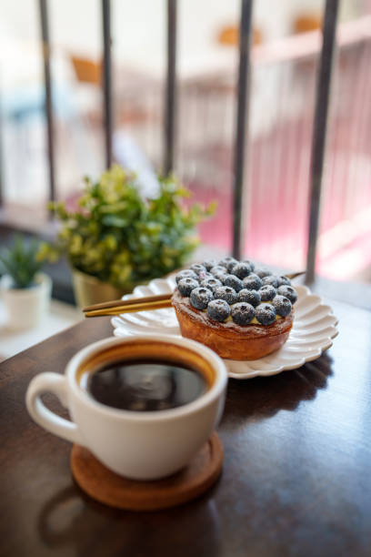 A cup of black coffee paired with a delicious blueberry tart on a white plate, placed on a wooden coaster. The cozy cafe setting includes indoor plants and a blurred view of a balcony.