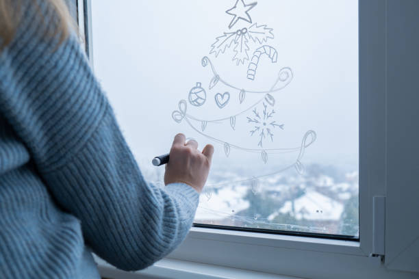 istock Creative enthusiastic unrecognizable woman in sweater paint window with white marker. Christmas tree decoration on big clear window. Snowy winter outside 2185338206