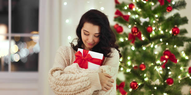 istock Woman in Cozy Sweater Embracing Christmas Gift by Decorated Tree 2185338355