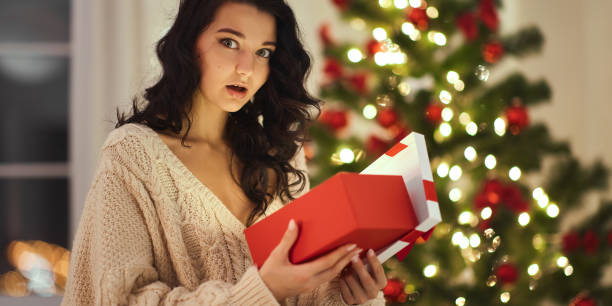istock Surprised Woman Opening Christmas Gift by Decorated Tree 2185339691