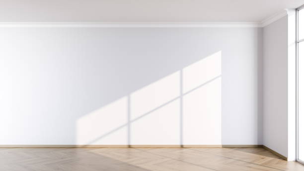 istock spacious, empty room features a light-colored wall and wooden floor. Bright natural light streams in through large windows, creating distinct shadows across the floor 2185355484