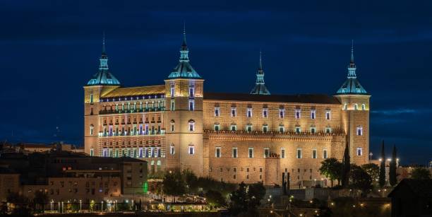 Aerial night view of the Alcázar of Toledo illuminated above the historic city of Toledo in central Spain. Warm city lights trace streets and medieval buildings surrounding the fortress, creating a striking contrast between light and shadow that highlights Spanish history, medieval architecture, and the atmospheric beauty of this UNESCO World Heritage city after dark. Castilla–La Mancha, central Spain, Europe.