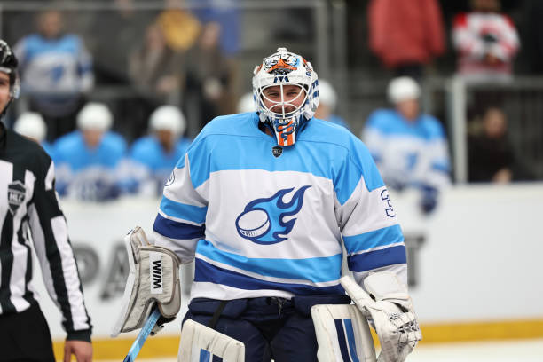 A blue team goalie in full protective gear stands focused, ready to defend the goal, with teammates and a referee visible in the background.
