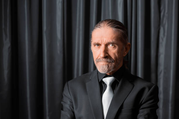 Elderly man wearing a black suit and white tie, with a stern expression, posing in front of dark curtains for a formal portrait.