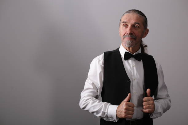 Elderly man wearing a bow tie and vest, confidently posing with thumbs up, against a plain gray studio background.