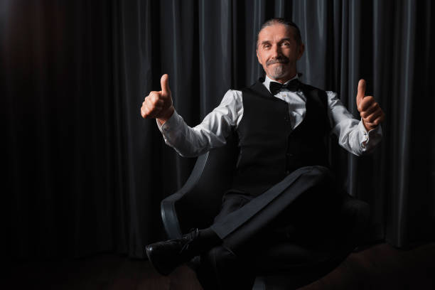 Stylish elderly man in formal attire sitting in a chair, smiling confidently, and giving a double thumbs up against a dark backdrop.
