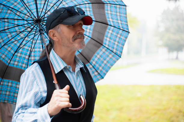 A senior businessman is using his mobile phone outside on a rainy day, looking at the screen and holding an umbrella