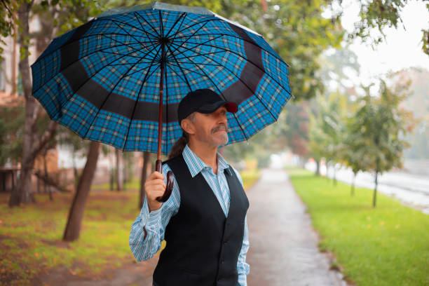 Mature man in a cap and vest holds a blue plaid umbrella while strolling along a rainy park path lined with trees.