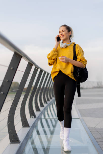 istock Cheerful woman strolls along a modern bridge, chatting on her phone while enjoying the serene cityscape at dusk by the water 2185379815