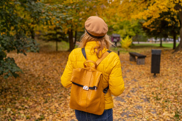 istock Back view of a woman with a yellow backpack walking through a leafy autumn path in fall. 2185386454